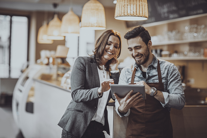 A man and a woman in a coffee shop working and looking at a tablet together